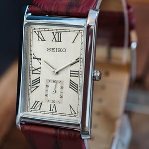 Puede incluir: Un reloj de pulsera Seiko rectangular con esfera plateada y números romanos. La correa es de cuero burdeos. La esfera muestra el nombre de la marca y un pequeño dial de segundos.