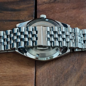 May include: Close-up of a stainless steel wristwatch with a silver-toned metal band. The watch face is visible, with the text "3309-6820 14". The watch is set against a dark wooden surface.