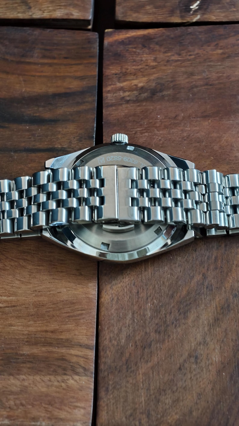 May include: Close-up of a stainless steel wristwatch with a silver-toned metal band. The watch face is visible, with the text "3309-6820 14". The watch is set against a dark wooden surface.