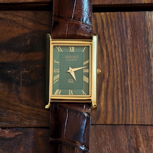 May include: A gold-coloured rectangular watch with a green face and a brown leather strap. The watch face displays Roman numerals and the Seiko logo. The strap is dark brown, textured leather. The watch is on a wooden surface.