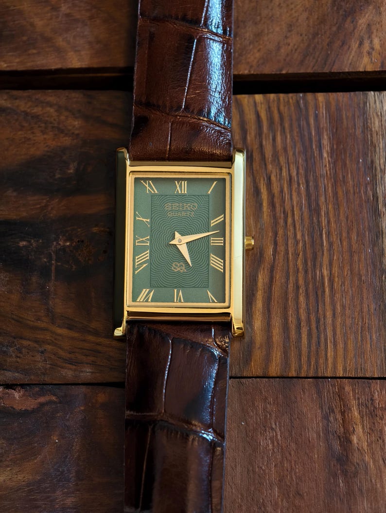 May include: A gold-coloured rectangular watch with a green face and a brown leather strap. The watch face displays Roman numerals and the Seiko logo. The strap is dark brown, textured leather. The watch is on a wooden surface.