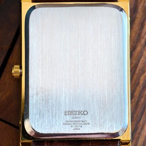 May include: A gold-coloured rectangular Seiko quartz watch with a brown leather strap. The watch back is stainless steel and features the Seiko logo and text indicating it is water-resistant. Made in Japan.