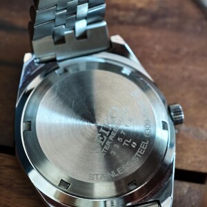 May include: Close-up of a stainless steel watch with a silver band. The watch face is visible, with the brand name "Seiko" and "Water Resistant" printed on it. The band has a woven design.