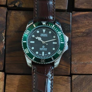 K&ouml;nnte beinhalten: Eine Seiko Automatic Submariner Uhr mit schwarzem Zifferblatt und gr&uuml;ner L&uuml;nette. Die Uhr hat ein braunes Lederarmband. Das Zifferblatt zeigt den Markennamen und weitere Details. Eine klassische Uhr.