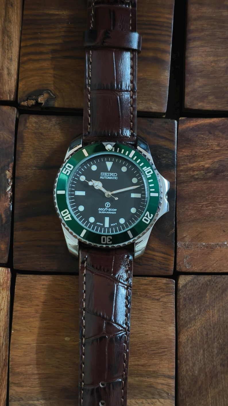 K&ouml;nnte beinhalten: Eine Seiko Automatic Submariner Uhr mit schwarzem Zifferblatt und gr&uuml;ner L&uuml;nette. Die Uhr hat ein braunes Lederarmband. Das Zifferblatt zeigt den Markennamen und weitere Details. Eine klassische Uhr.