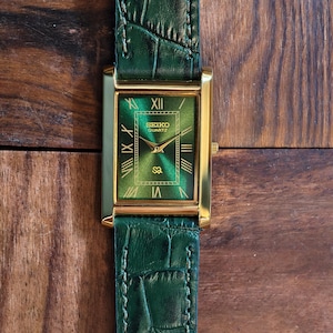 May include: A rectangular gold-toned Seiko Quartz watch with a green face and Roman numeral hour markers. The watch has a green leather band with a crocodile pattern. The watch is displayed on a wooden surface.