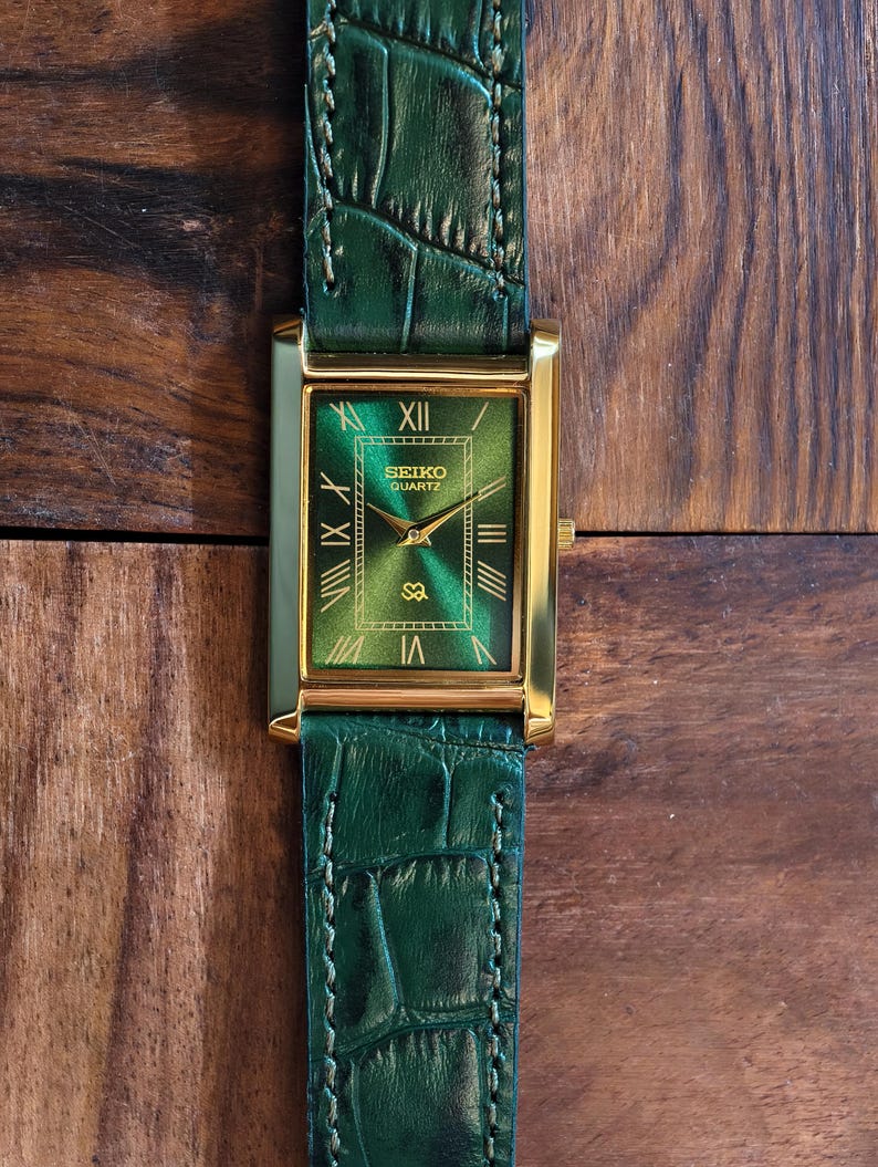 May include: A rectangular gold-toned Seiko Quartz watch with a green face and Roman numeral hour markers. The watch has a green leather band with a crocodile pattern. The watch is displayed on a wooden surface.