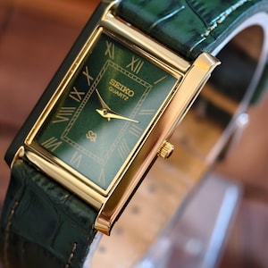 May include: A rectangular gold-toned Seiko Quartz watch with a green face and gold hands. The watch features Roman numerals and a green leather strap. The watch face is approximately 2.5 cm wide.
