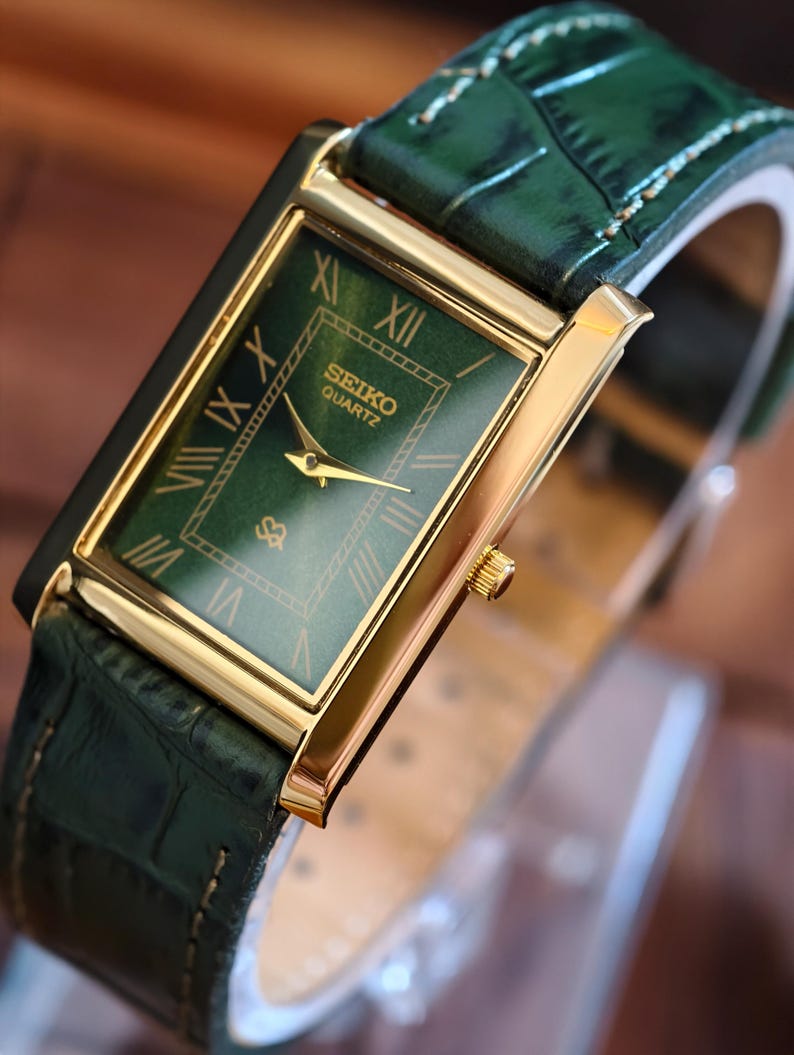 May include: A rectangular gold-toned Seiko Quartz watch with a green face and gold hands. The watch features Roman numerals and a green leather band. The watch face is approximately 1 inch wide.
