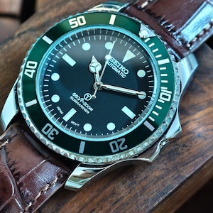 K&ouml;nnte beinhalten: Seiko Automatic Submariner Uhr aus Edelstahl mit schwarzem Zifferblatt und gr&uuml;ner L&uuml;nette. Das Armband ist aus braunem Leder und die Uhr liegt auf einem Holzblock. Das Zifferblatt zeigt die Uhrzeit und weitere Informationen.