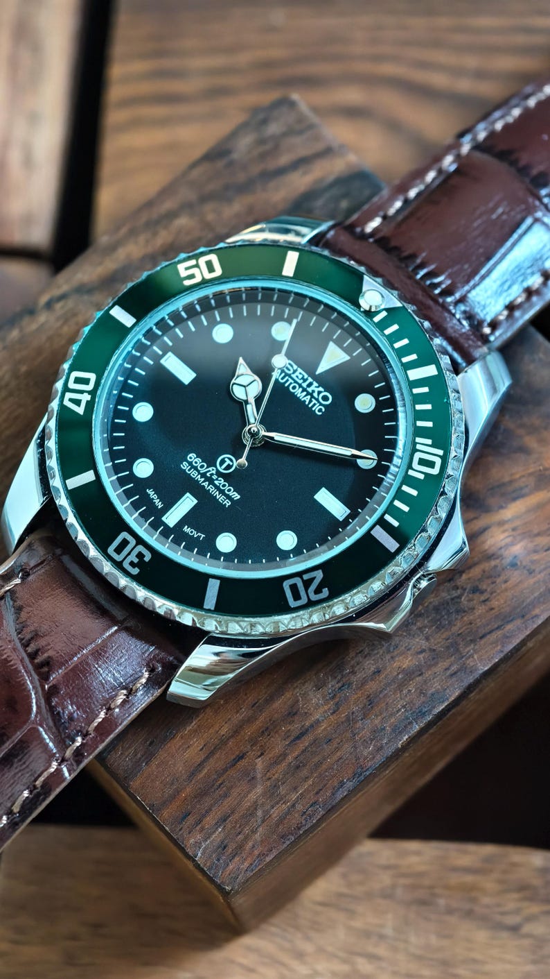 K&ouml;nnte beinhalten: Seiko Automatic Submariner Uhr aus Edelstahl mit schwarzem Zifferblatt und gr&uuml;ner L&uuml;nette. Das Armband ist aus braunem Leder und die Uhr liegt auf einem Holzblock. Das Zifferblatt zeigt die Uhrzeit und weitere Informationen.