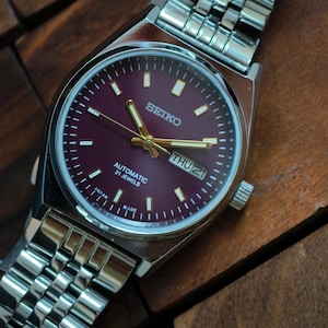 May include: A stainless steel Seiko automatic watch with a burgundy dial. The watch features gold hands and markers, a day-date display, and a silver link bracelet. The watch face displays "SEIKO", "AUTOMATIC 21 JEWELS", and "MADE IN JAPAN".