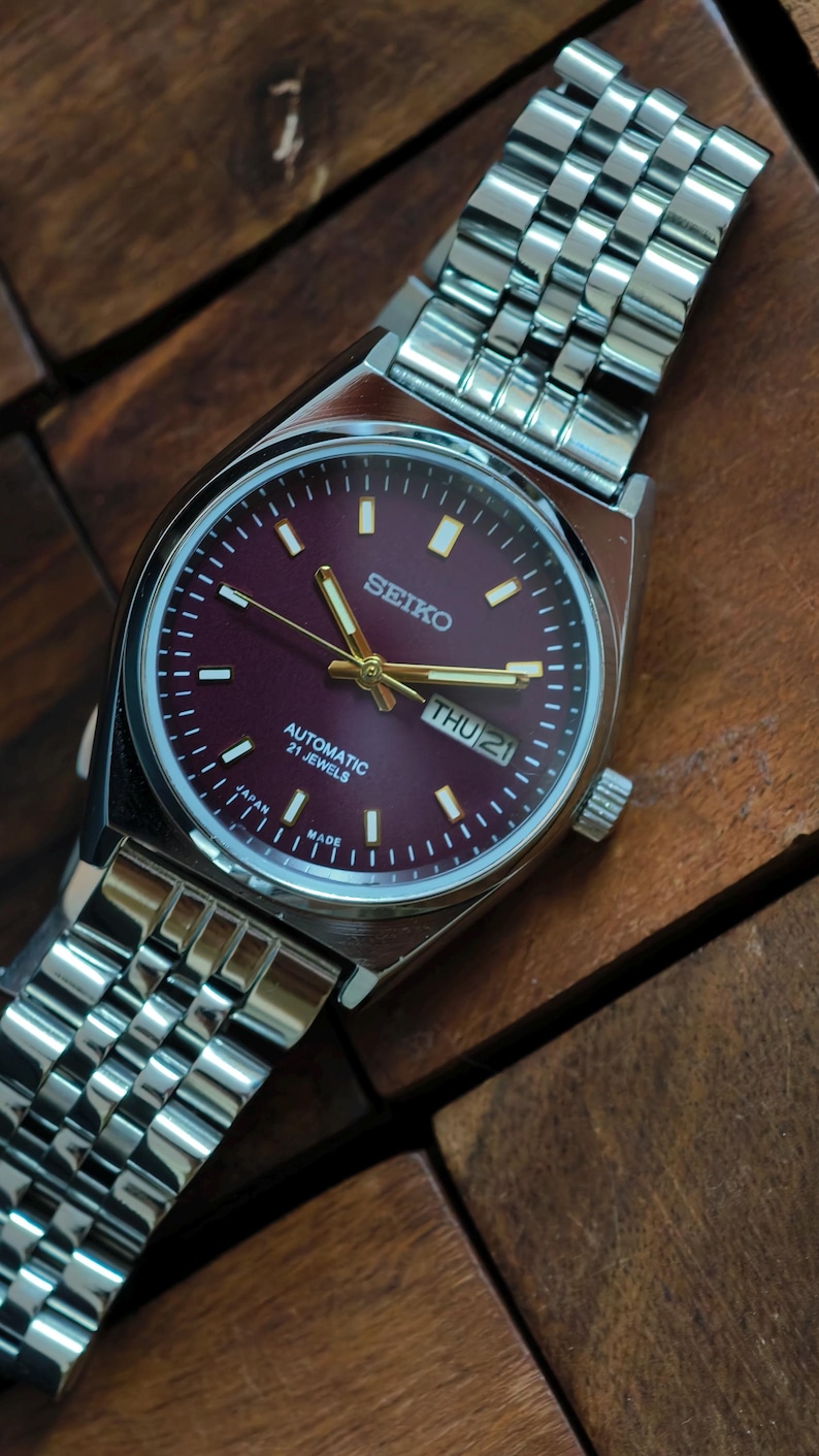 May include: A stainless steel Seiko automatic watch with a burgundy dial. The watch features gold hands and markers, a day-date display, and a silver link bracelet. The watch face displays "SEIKO", "AUTOMATIC 21 JEWELS", and "MADE IN JAPAN".