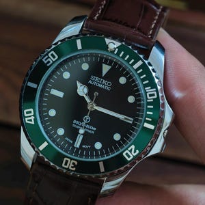 Op de afbeelding: Een roestvrijstalen Seiko automatisch horloge met een zwarte wijzerplaat en een groene lunette. Het horloge heeft een bruine leren band. De wijzerplaat toont de tekst "SEIKO AUTOMATIC" en "SUBMARINER".
