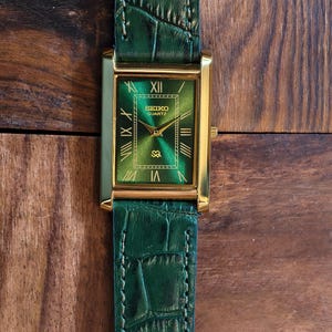 May include: A rectangular Seiko Quartz watch with a gold-tone case and a green dial. The watch features Roman numeral hour markers and gold hands. The green leather band has a crocodile pattern.