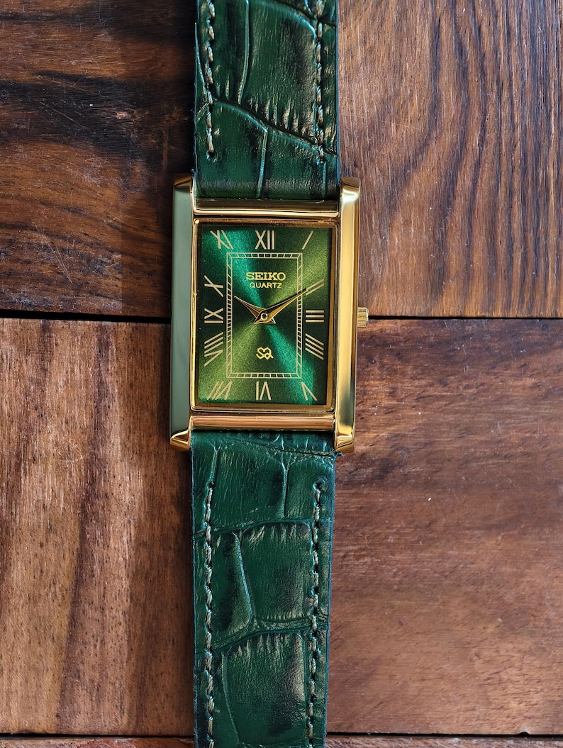 May include: A rectangular Seiko Quartz watch with a gold-tone case and a green dial. The watch features Roman numeral hour markers and gold hands. The green leather band has a crocodile pattern.