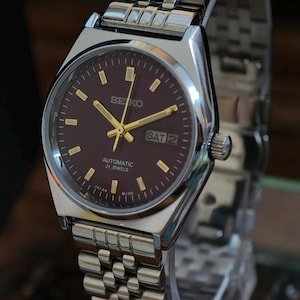 May include: A silver-toned Seiko automatic wristwatch with a brown dial and gold-colored hands and markers. The watch displays the day and date. The watchband is made of silver-toned metal links. The watch is marked "Japan Made".