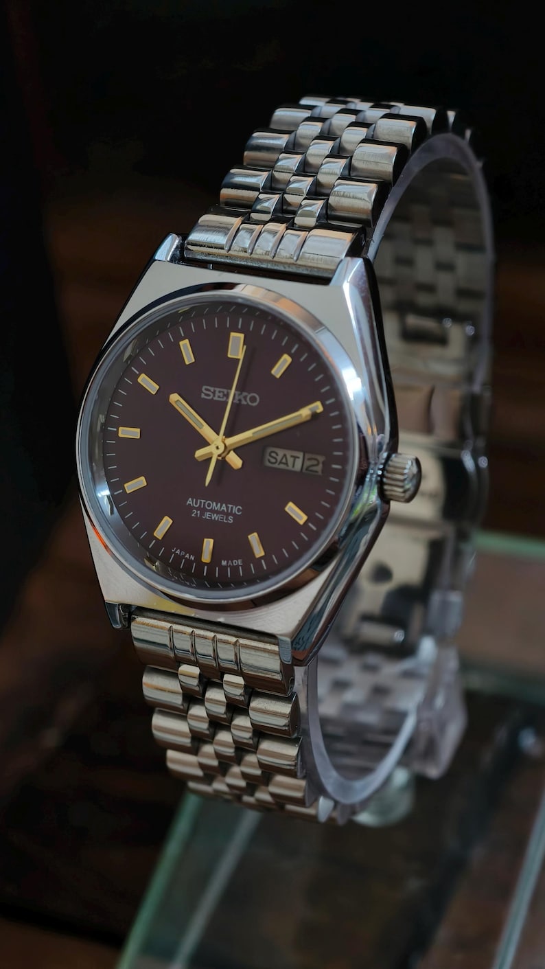 May include: A silver-toned Seiko automatic wristwatch with a brown dial and gold-colored hands and markers. The watch displays the day and date. The watchband is made of silver-toned metal links. The watch is marked "Japan Made".