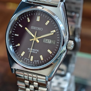 May include: A silver-toned Seiko automatic wristwatch with a dark brown face. The watch features gold hands and markers, a day-date display, and a stainless steel band. The watch is marked "Japan Made".
