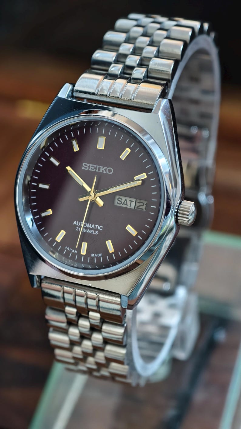 May include: A silver-toned Seiko automatic wristwatch with a dark brown face. The watch features gold hands and markers, a day-date display, and a stainless steel band. The watch is marked "Japan Made".