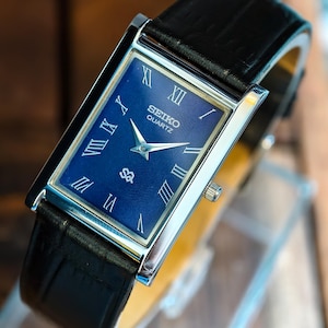 May include: A rectangular Seiko Quartz watch with a navy blue face and silver hands. The watch has Roman numeral hour markers and a black leather strap. The watch face has the Seiko Quartz logo and a small emblem.