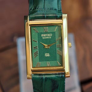 May include: A rectangular gold-tone Seiko Quartz watch with a green face and green leather strap. The watch face features gold Roman numerals and hands. The strap is a textured green leather.