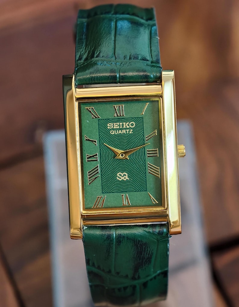 May include: A rectangular gold-tone Seiko Quartz watch with a green face and green leather strap. The watch face features gold Roman numerals and hands. The strap is a textured green leather.