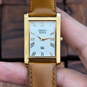 Peut inclure: Une montre Seiko Quartz dorée de forme rectangulaire avec un cadran blanc et des chiffres romains. Elle est dotée d'un bracelet en cuir marron. Le cadran affiche le logo Seiko Quartz et un petit symbole.