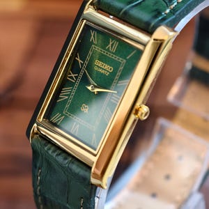 May include: A rectangular Seiko Quartz watch with a green dial and gold-tone case. The watch features Roman numeral hour markers and a green leather strap with a crocodile pattern. The watch has a gold crown and hands.