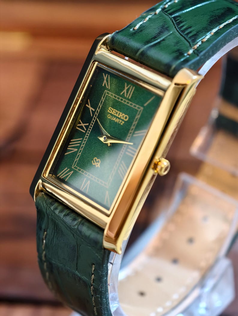 May include: A rectangular Seiko Quartz watch with a green dial and gold-tone case. The watch features Roman numeral hour markers and a green leather strap with a crocodile pattern. The watch has a gold crown and hands.