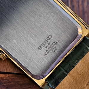 May include: Close-up of a vintage Seiko quartz watch. The watch features a gold-toned rectangular case with a stainless steel back. The back is engraved with "SEIKO QUARTZ WATER RESISTANT STAINLESS STEEL BACK SO.262714 JAPAN". A green leather strap is attached.