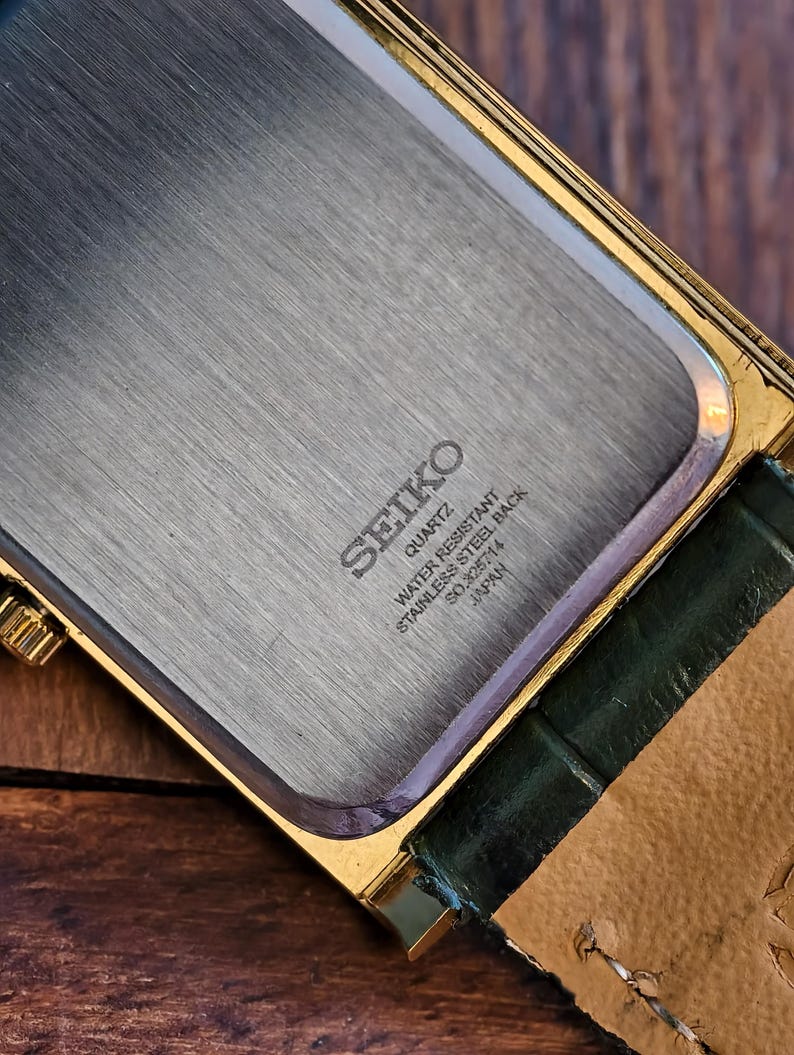 May include: Close-up of a vintage Seiko quartz watch. The watch features a gold-toned rectangular case with a stainless steel back. The back is engraved with "SEIKO QUARTZ WATER RESISTANT STAINLESS STEEL BACK SO.262714 JAPAN". A green leather strap is attached.
