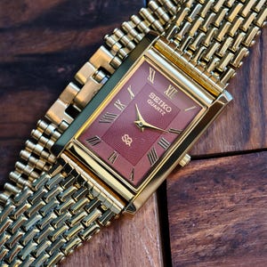 May include: A gold Seiko Quartz watch with a rectangular maroon face and gold Roman numerals. The watch has a gold mesh strap and is displayed on a wooden surface. The watch face has the Seiko Quartz logo.