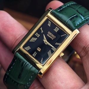 Könnte beinhalten: Rechteckige Seiko Quartz-Uhr mit schwarzem Zifferblatt, goldfarbenem Gehäuse und grünem Lederarmband. Die Uhr hat goldene römische Ziffern und Zeiger. Klassisches Design.