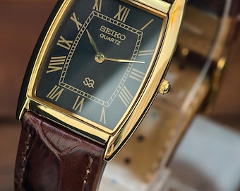 May include: A rectangular Seiko Quartz watch with a gold-tone case and a dark face featuring gold Roman numerals. The watch has a brown leather band. The watch face displays the Seiko Quartz logo.