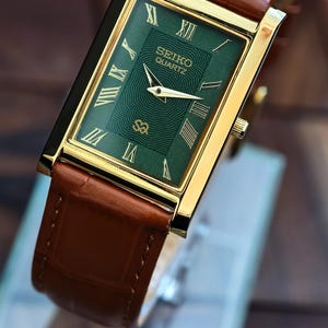 Könnte beinhalten: Rechteckige Seiko Quartz-Uhr mit grünem Zifferblatt und goldfarbenem Gehäuse. Die Uhr hat römische Ziffern und ein braunes Lederarmband. Das Zifferblatt zeigt das Seiko Quartz-Logo. Klassisches Design.