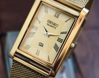 Reloj de cuarzo Seiko vintage: esfera dorada con números romanos y diseño delgado.