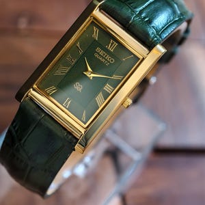 May include: A rectangular Seiko Quartz watch with a gold-tone case and a green dial with Roman numerals. The watch has a green leather strap. The watch face displays the Seiko Quartz logo. A classic timepiece.