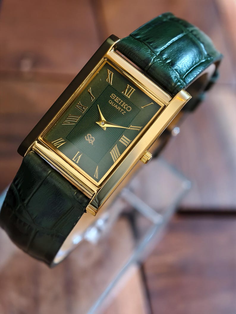 May include: A rectangular Seiko Quartz watch with a gold-tone case and a green dial with Roman numerals. The watch has a green leather strap. The watch face displays the Seiko Quartz logo. A classic timepiece.