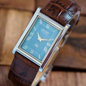 May include: A rectangular Seiko watch with a light blue face and gold hands, featuring Roman numerals. The watch has a silver case and a brown leather strap with a crocodile pattern.