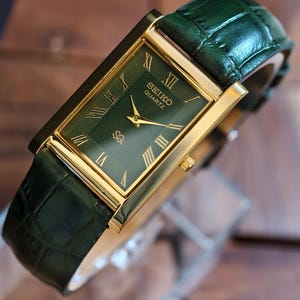 May include: A rectangular Seiko Quartz watch with a gold-tone case and a green dial. The watch features gold hands and Roman numeral hour markers. The watch has a green leather strap with a crocodile pattern.