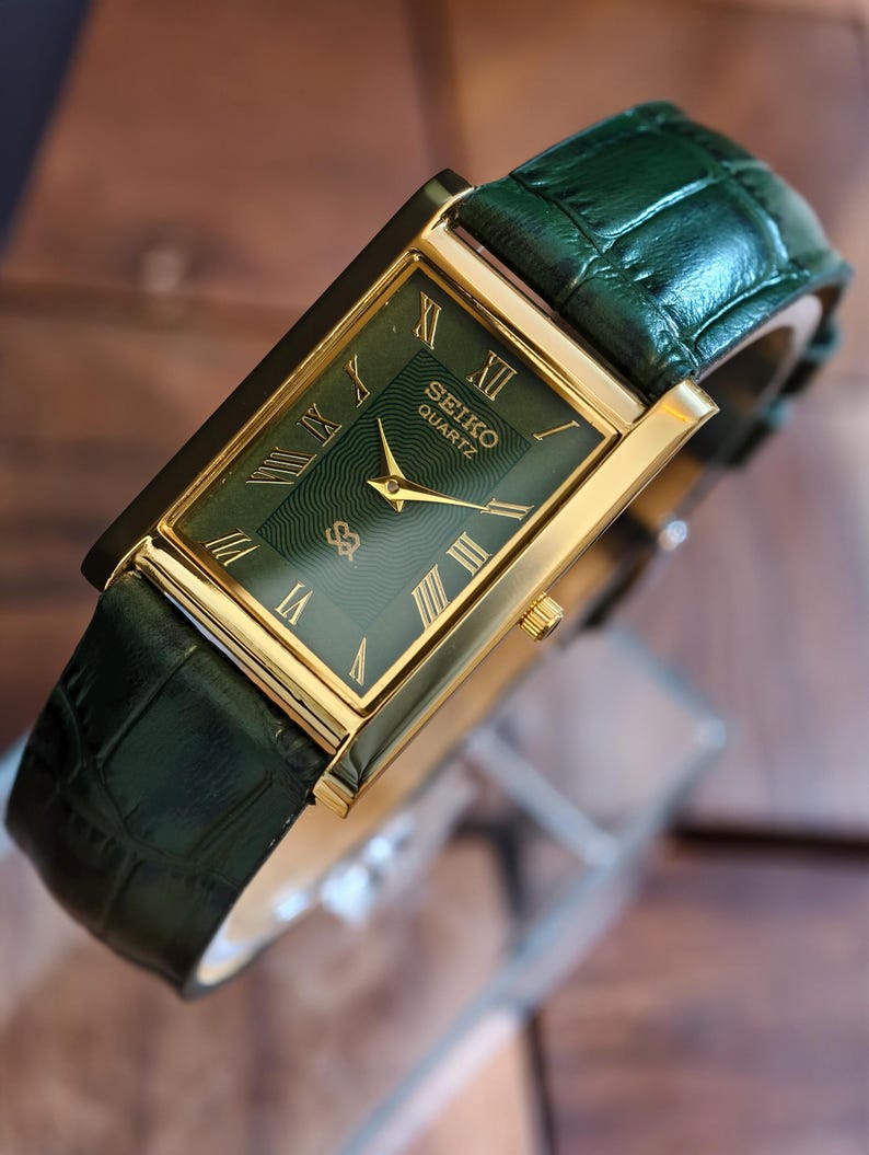 May include: A rectangular Seiko Quartz watch with a gold-tone case and a green dial. The watch features gold hands and Roman numeral hour markers. The watch has a green leather strap with a crocodile pattern.