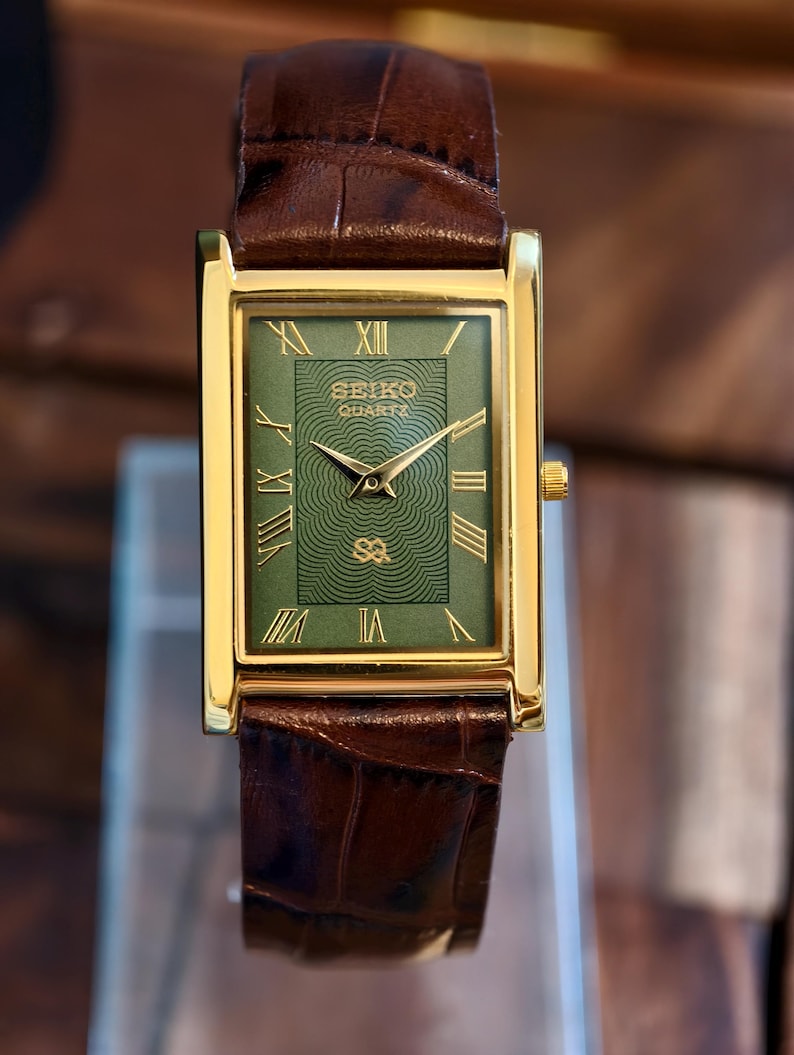 May include: A gold-coloured rectangular Seiko Quartz watch with a green face and brown leather strap. The watch face has gold Roman numerals and hands. The strap is a dark brown, textured leather.