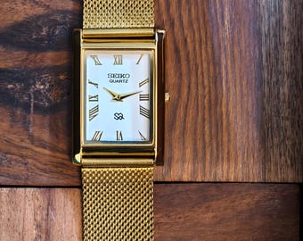 Può includere: Orologio Seiko Quartz dorato con quadrante rettangolare e cinturino a maglia. Il quadrante bianco dell'orologio presenta numeri romani neri e lancette dorate. L'orologio è appoggiato su una superficie di legno.
