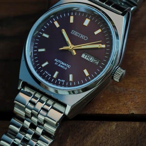 May include: A silver Seiko automatic wristwatch with a dark purple dial and gold hands. The watch face displays the day and date, with the text "SEIKO", "AUTOMATIC 21 JEWELS", and "JAPAN MADE". The watch has a silver metal band.