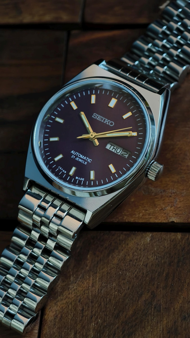 May include: A silver Seiko automatic wristwatch with a dark purple dial and gold hands. The watch face displays the day and date, with the text "SEIKO", "AUTOMATIC 21 JEWELS", and "JAPAN MADE". The watch has a silver metal band.