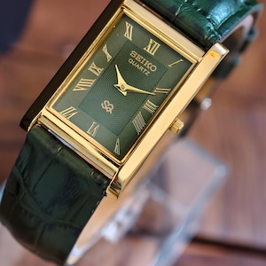 May include: A rectangular Seiko Quartz watch with a gold-tone case and a green dial. The watch features Roman numeral hour markers and a green leather strap. The watch face displays the Seiko Quartz logo.