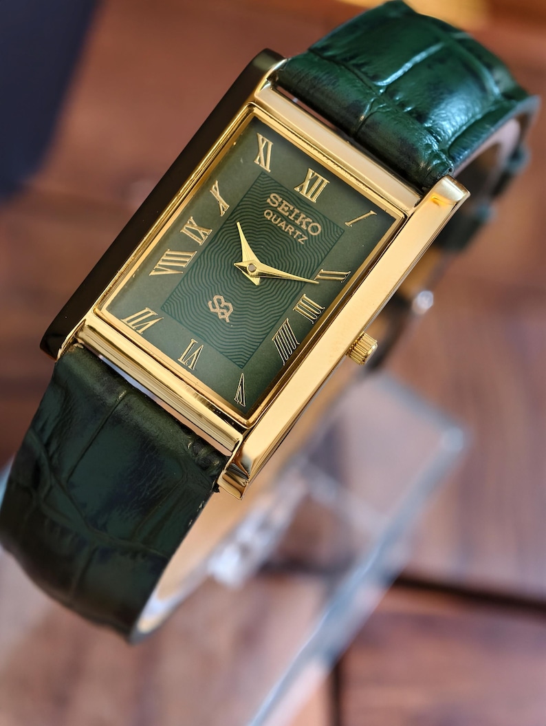 May include: A rectangular Seiko Quartz watch with a gold-tone case and a green dial. The watch features Roman numeral hour markers and a green leather strap. The watch face displays the Seiko Quartz logo.