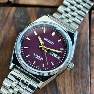 May include: A silver-toned Seiko wristwatch with a burgundy face and a silver band. The watch face displays the day and date, with gold-colored hands and markers. The watch is labeled "Automatic 21 Jewels" and "Japan Made".