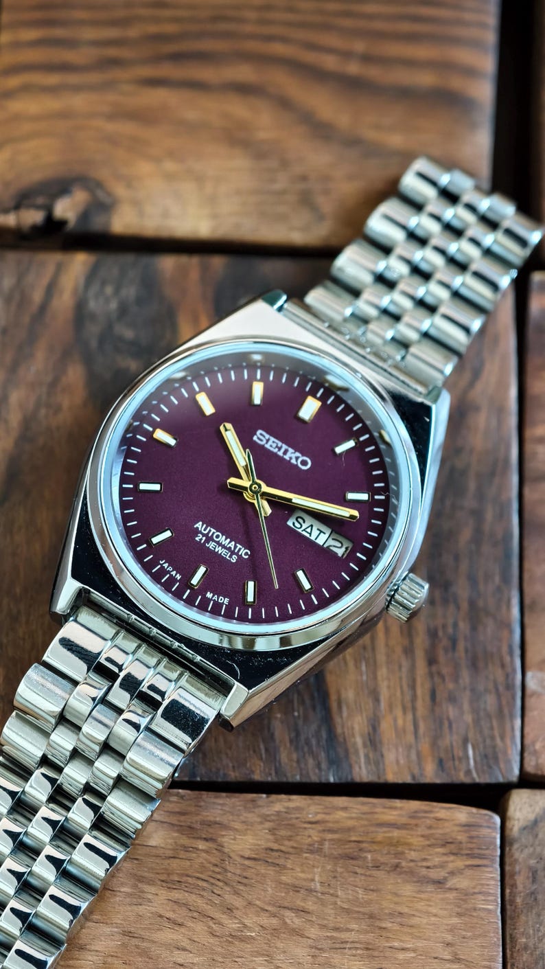 May include: A silver-toned Seiko wristwatch with a burgundy face and a silver band. The watch face displays the day and date, with gold-colored hands and markers. The watch is labeled "Automatic 21 Jewels" and "Japan Made".